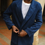 Close-up of a man buttoning a navy blue corduroy jacket worn over a white shirt with navy stripes. The jacket features wide lapels and matching corduroy pants.