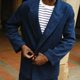 Close-up of a man buttoning a navy blue corduroy jacket worn over a white shirt with navy stripes. The jacket features wide lapels and matching corduroy pants.