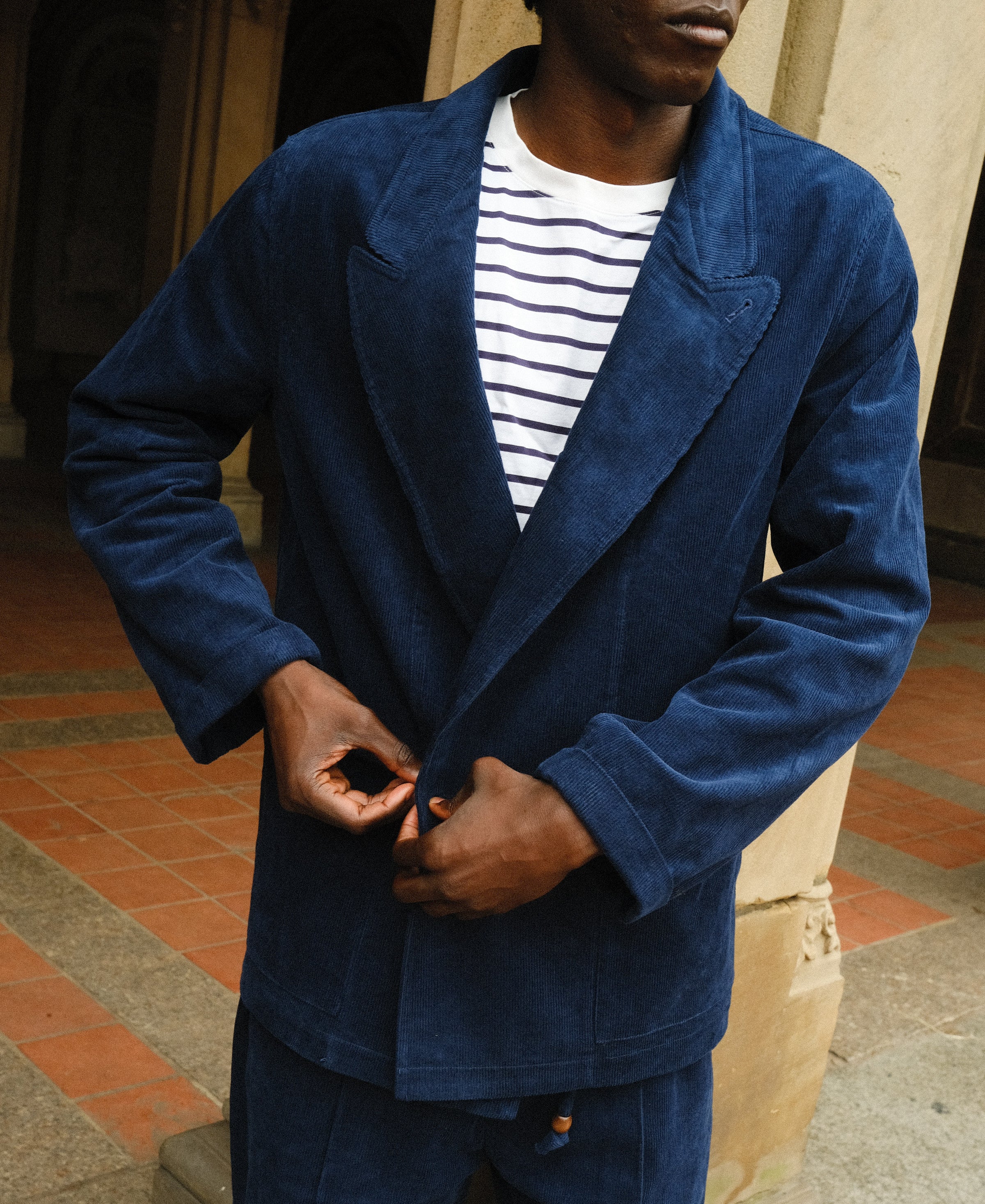Close-up of a man buttoning a navy blue corduroy jacket worn over a white shirt with navy stripes. The jacket features wide lapels and matching corduroy pants.