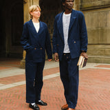 Two people wearing matching navy blue corduroy suits. The person on the left pairs theirs with a white top, white socks, and black loafers, while the person on the right wears a striped shirt and brown loafers, holding a stack of books.