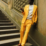 A man standing on stone steps, wearing a yellow corduroy suit with a relaxed double-breasted jacket and matching trousers. He pairs it with a white crewneck top and brown leather loafers. He is tossing a red apple in the air and leaning casually against a textured wall.