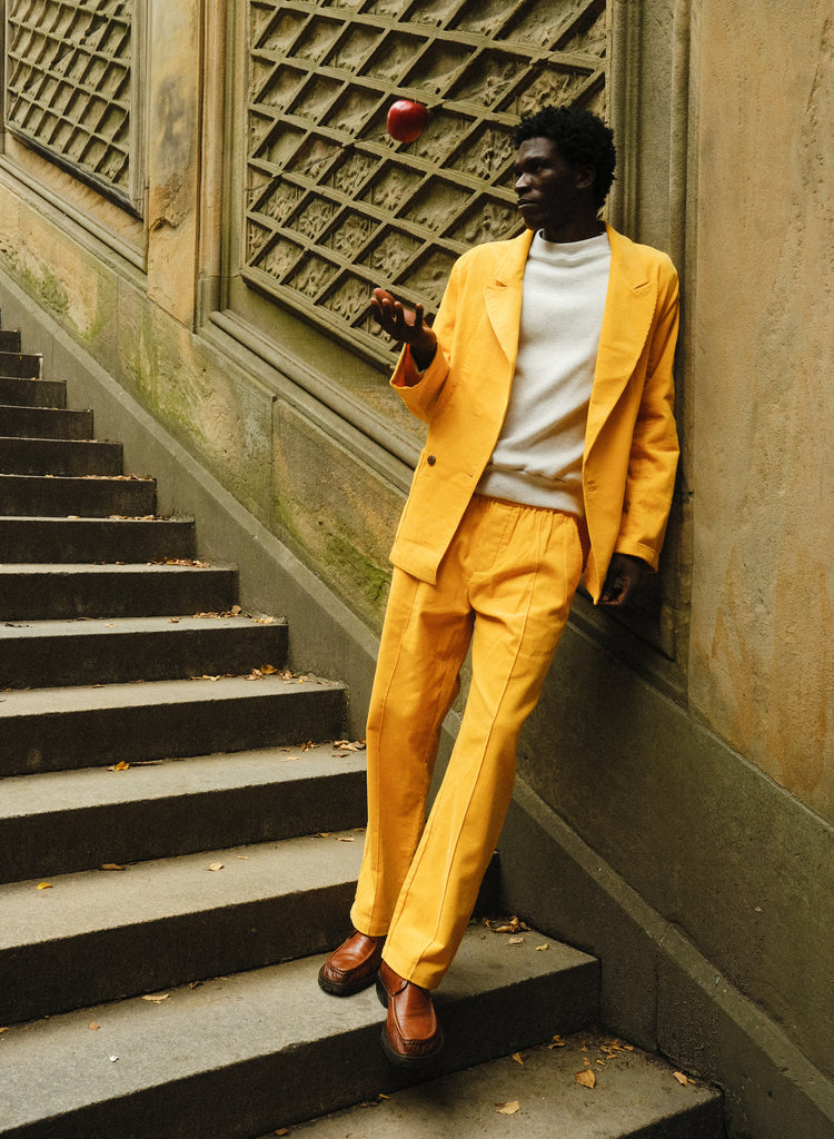 A man standing on stone steps, wearing a yellow corduroy suit with a relaxed double-breasted jacket and matching trousers. He pairs it with a white crewneck top and brown leather loafers. He is tossing a red apple in the air and leaning casually against a textured wall.