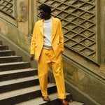 A man standing on stone steps wearing a yellow corduroy suit with a relaxed double-breasted jacket and matching trousers. He pairs it with a white crewneck top and brown leather loafers. 