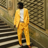 A man standing on stone steps wearing a yellow corduroy suit with a relaxed double-breasted jacket and matching trousers. He pairs it with a white crewneck top and brown leather loafers. 