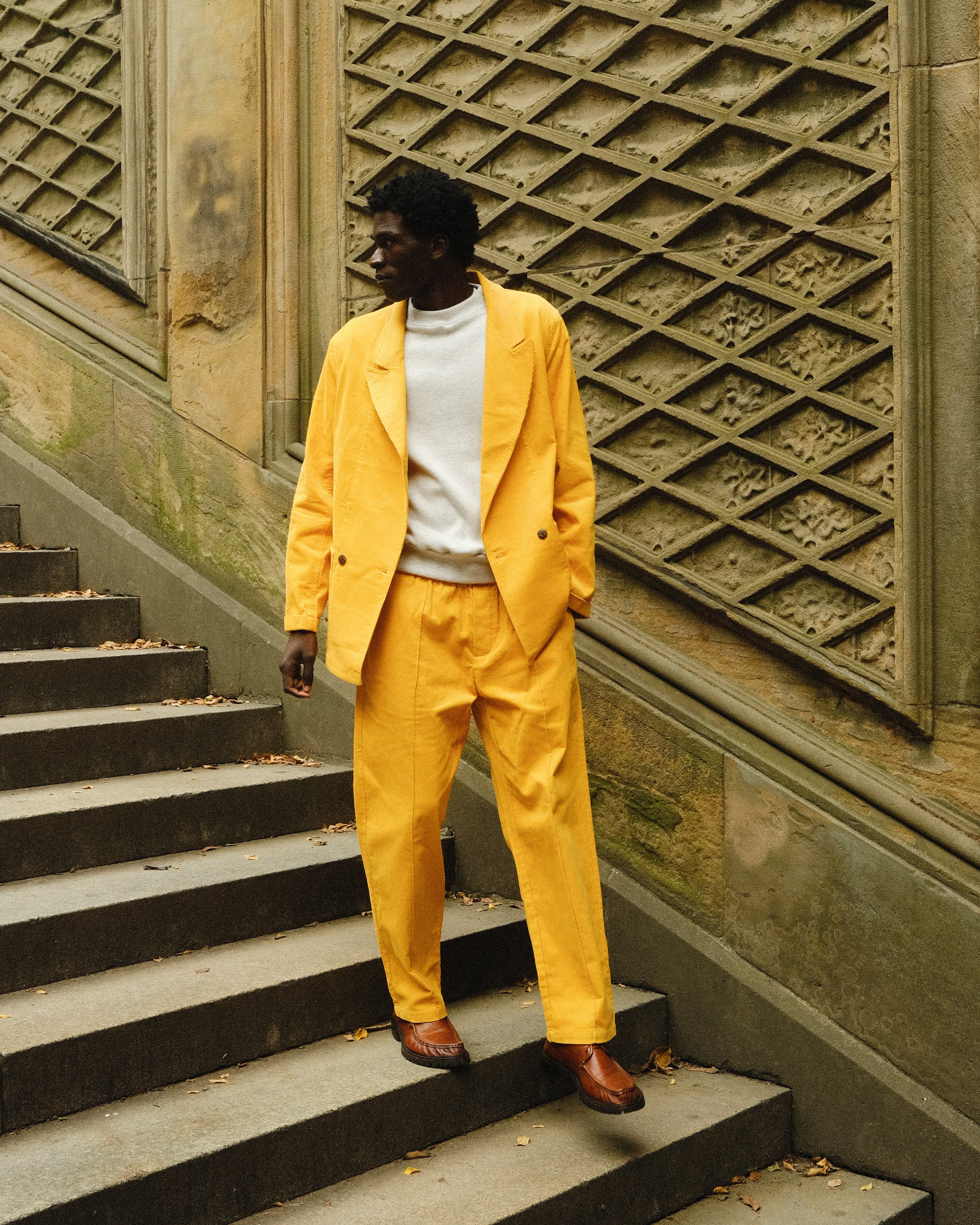A man standing on stone steps wearing a yellow corduroy suit with a relaxed double-breasted jacket and matching trousers. He pairs it with a white crewneck top and brown leather loafers. 