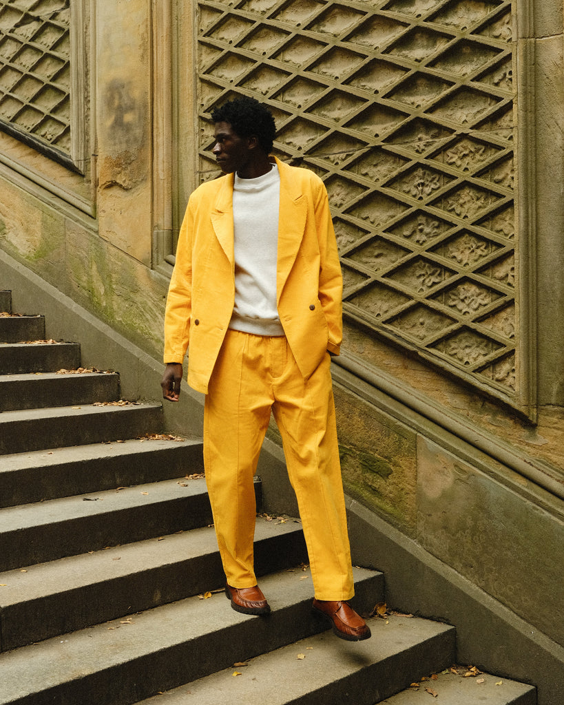 A man standing on stone steps wearing a yellow corduroy suit with a relaxed double-breasted jacket and matching trousers. He pairs it with a white crewneck top and brown leather loafers. 