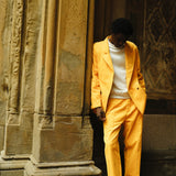 A man wearing a yellow corduroy suit with a relaxed double-breasted jacket and matching trousers. He pairs it with a white crewneck top and brown leather loafers. He is leaning casually against a textured wall.