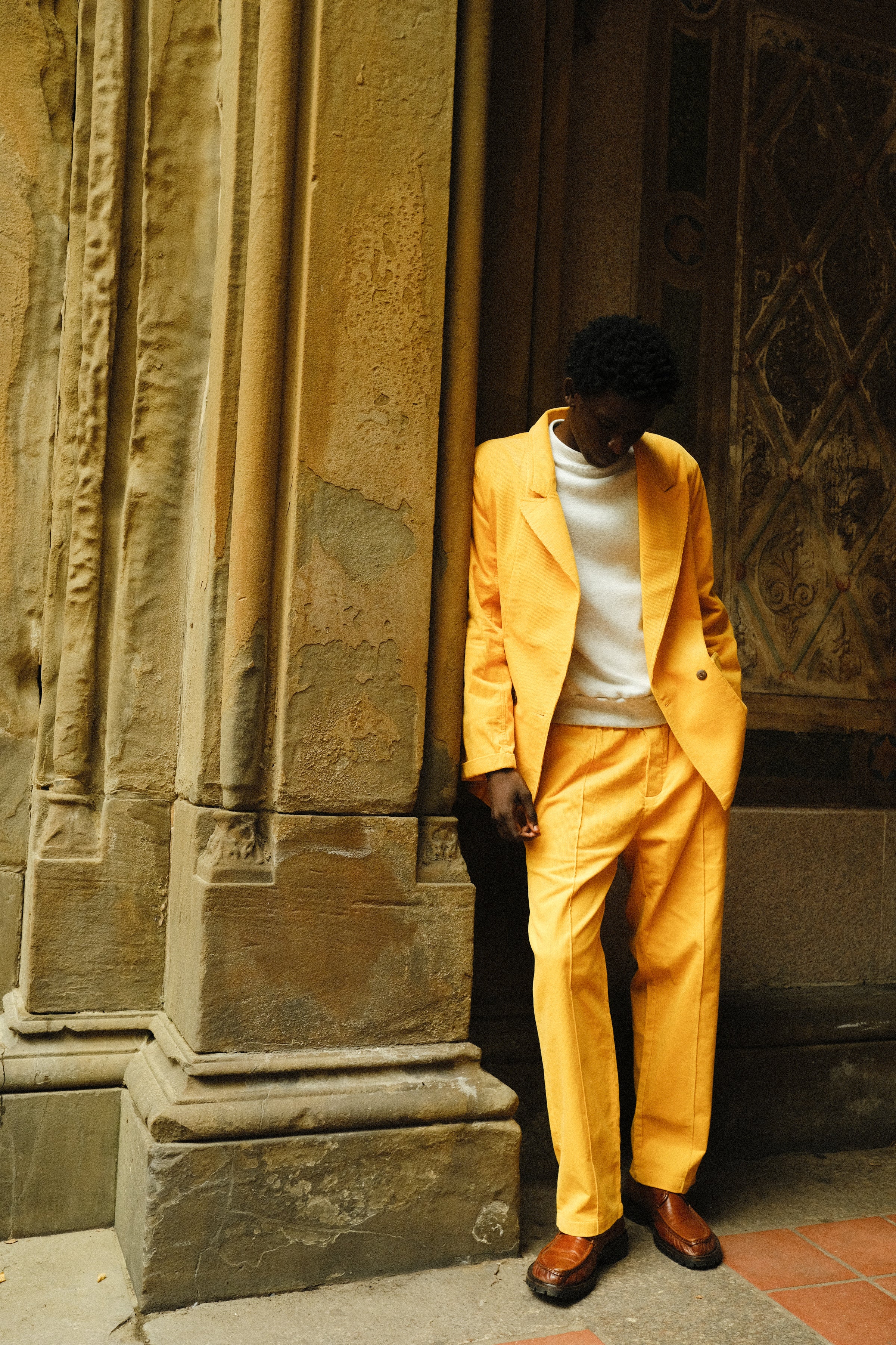 A man wearing a yellow corduroy suit with a relaxed double-breasted jacket and matching trousers. He pairs it with a white crewneck top and brown leather loafers. He is leaning casually against a textured wall.