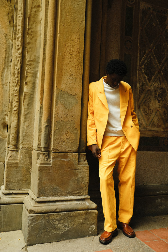A man wearing a yellow corduroy suit with a relaxed double-breasted jacket and matching trousers. He pairs it with a white crewneck top and brown leather loafers. He is leaning casually against a textured wall.