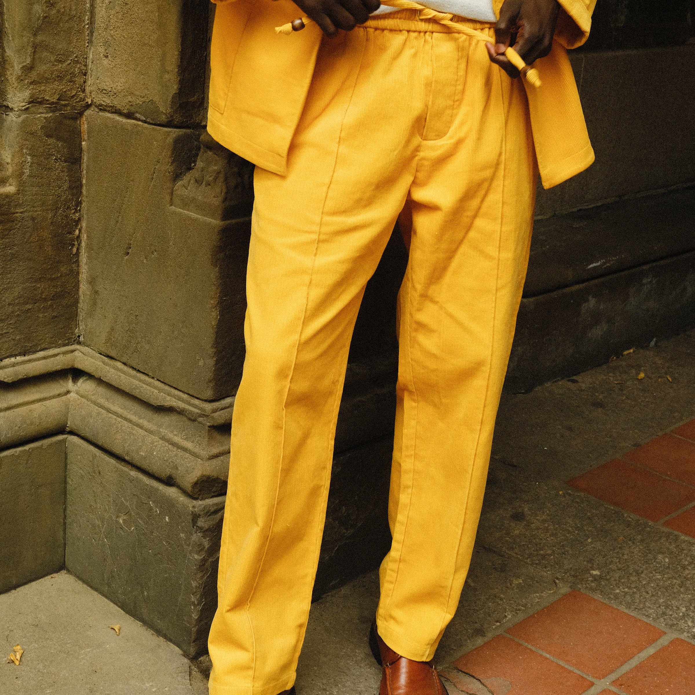 Close-up of a person wearing a bright yellow corduroy jacket and drawstring pants, paired with brown loafers and a light gray shirt.