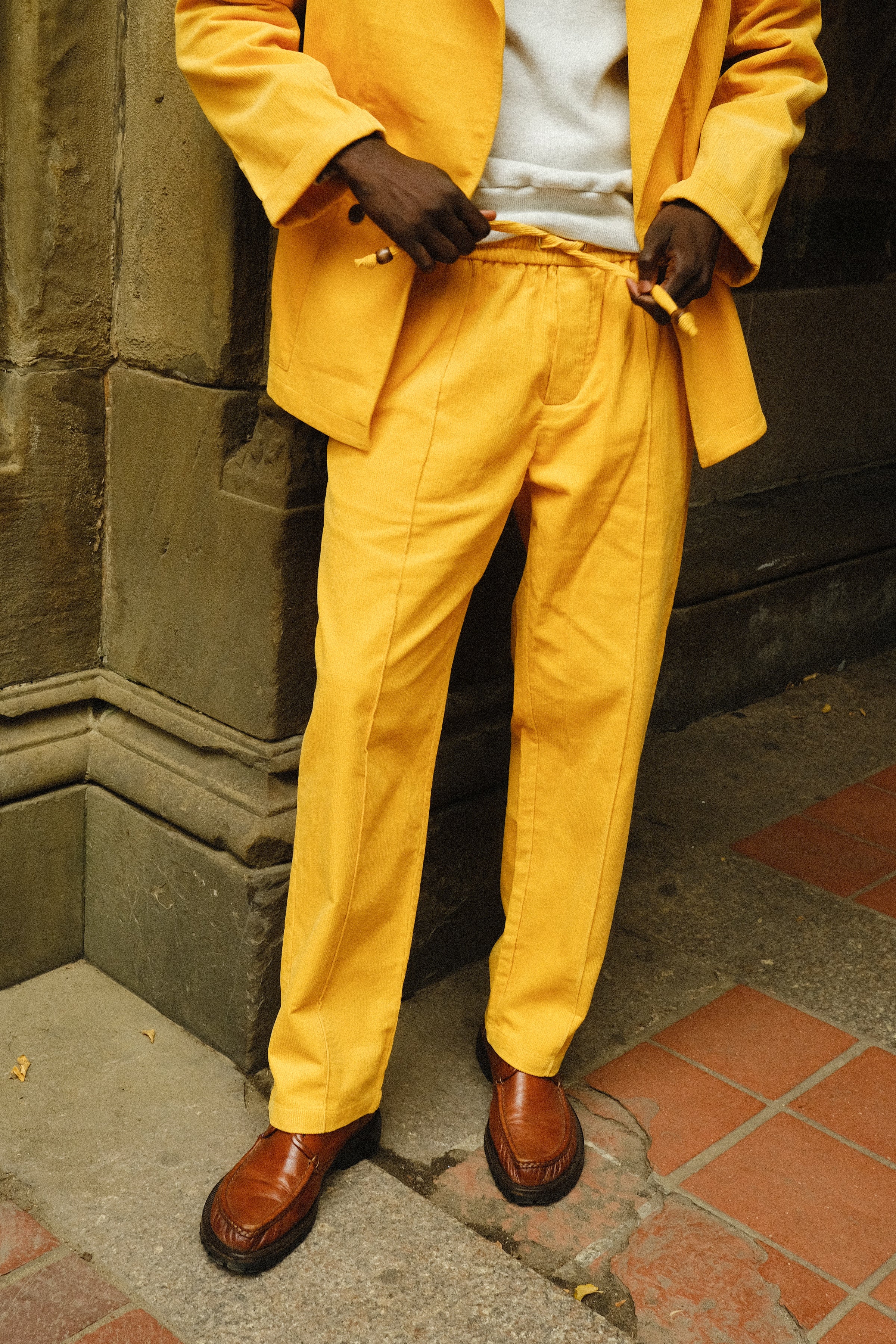 Close-up of a person wearing a bright yellow corduroy jacket and drawstring pants, paired with brown loafers and a light gray shirt.