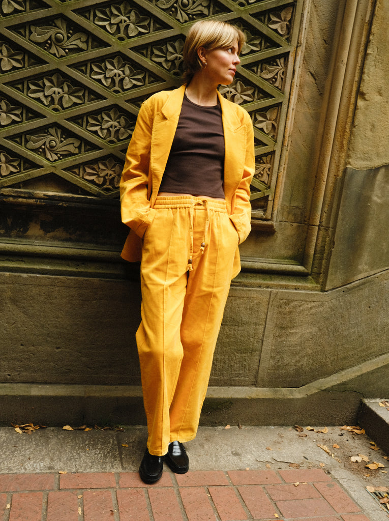 A woman stands against a wall, wearing a matching yellow corduroy set with a relaxed blazer and drawstring trousers. They pair the look with a fitted brown tank top and black loafers.