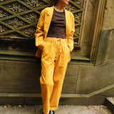 A woman stands against a wall, wearing a matching yellow corduroy set with a relaxed blazer and drawstring trousers. They pair the look with a fitted brown tank top and black loafers.