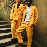 Two people standing on steps, both dressed in matching yellow corduroy suits. One wears the suit with a brown tank top and black loafers, while the other pairs it with a white crewneck and brown shoes. 