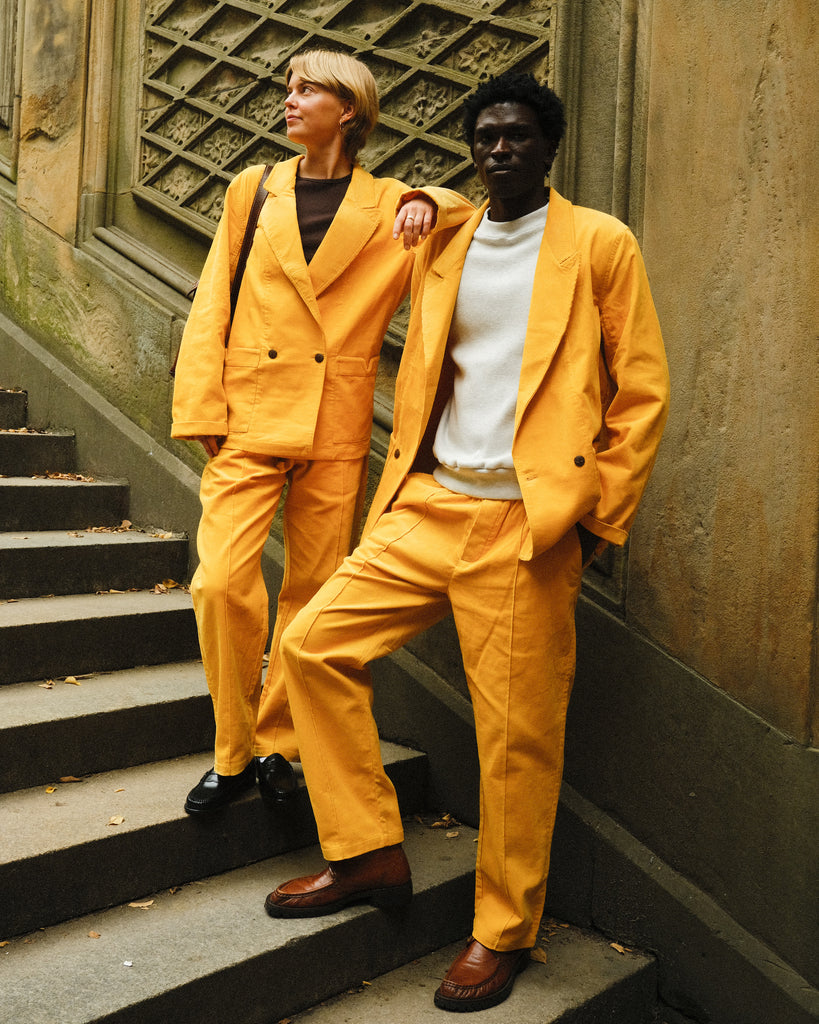 Two people standing on steps, both dressed in matching yellow corduroy suits. One wears the suit with a brown tank top and black loafers, while the other pairs it with a white crewneck and brown shoes. 