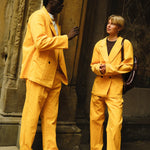 Two people wearing matching saffron yellow corduroy suits. The person on the left wears brown loafers and a light gray shirt, while the person on the right wears black shoes and a dark brown top with a backpack over one shoulder.