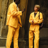Two people wearing matching saffron yellow corduroy suits. The person on the left wears brown loafers and a light gray shirt, while the person on the right wears black shoes and a dark brown top with a backpack over one shoulder.
