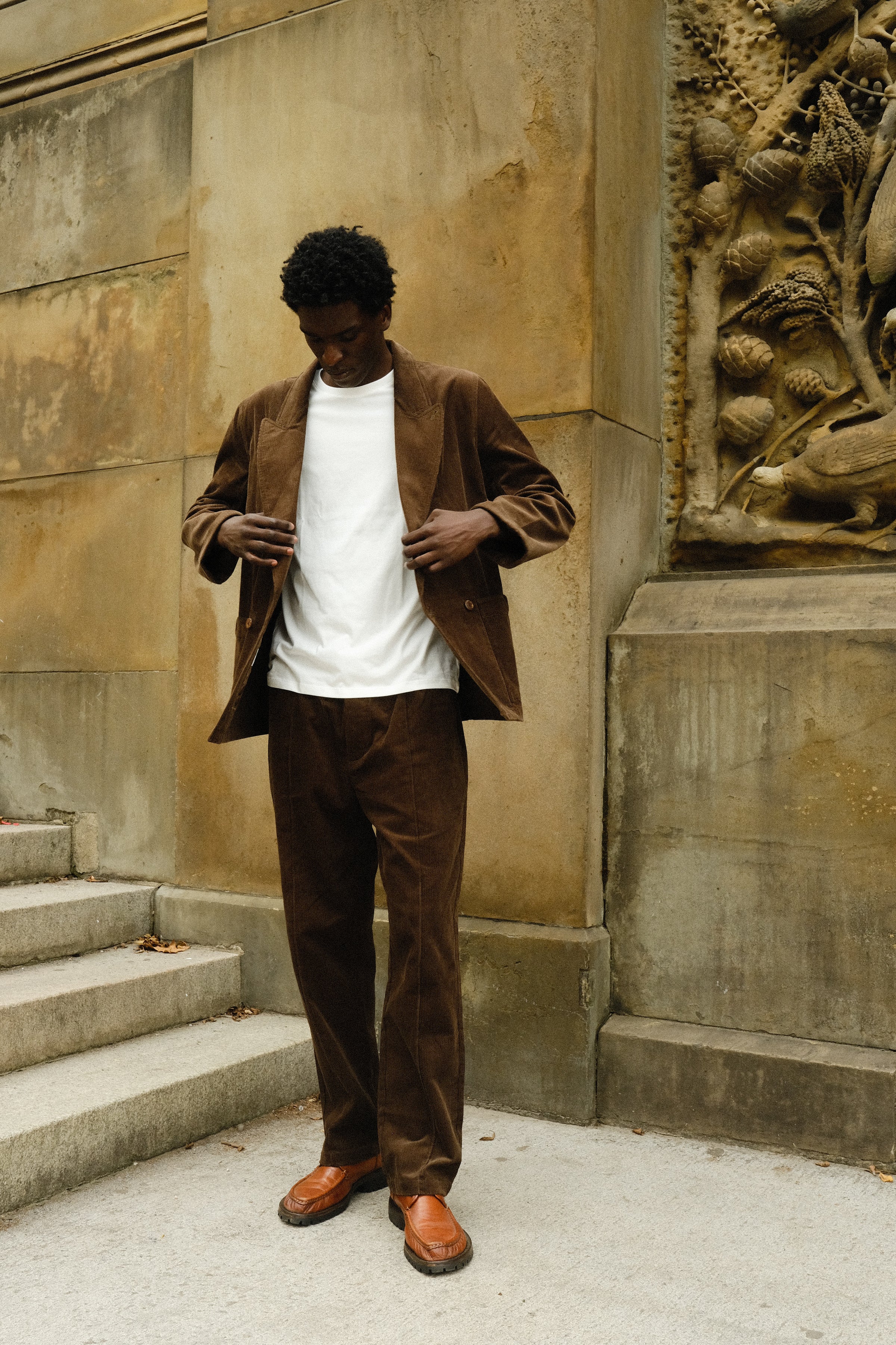 Man wearing a brown corduroy jacket and matching pants, paired with a plain white T-shirt and brown loafers.