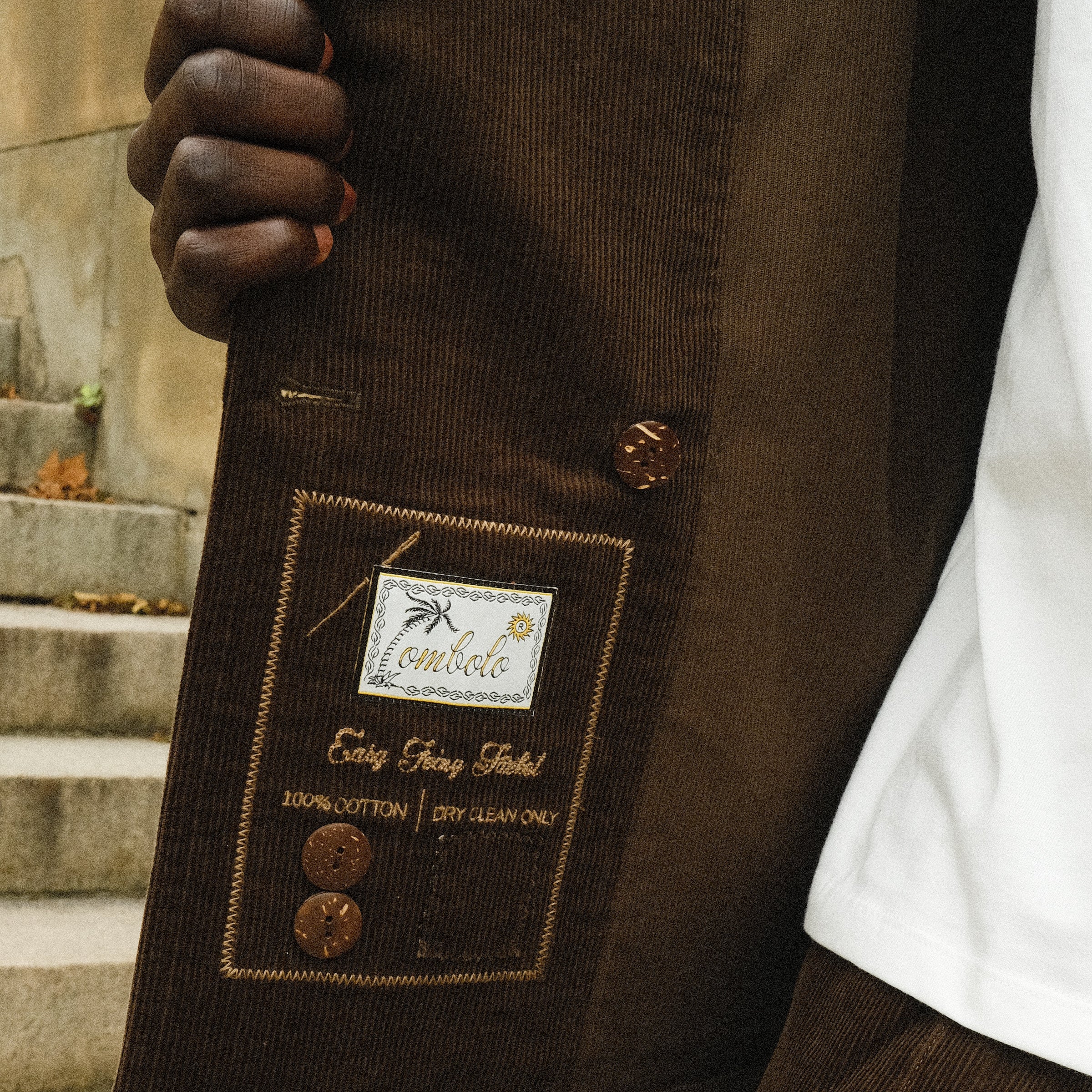 Close-up of the inside label on a brown corduroy jacket, showing embroidered branding that reads 'Tombolo' and 'Easy Going Jacket' with care instructions and two spare brown buttons attached.