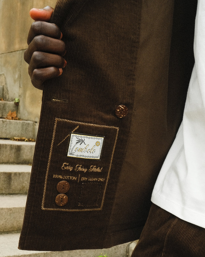 Close-up of the inside label on a brown corduroy jacket, showing embroidered branding that reads 'Tombolo' and 'Easy Going Jacket' with care instructions and two spare brown buttons attached.