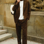 Man wearing a brown corduroy jacket and matching pants with a white T-shirt, brown loafers, and a white canvas tote bag slung over his shoulder.