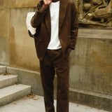 Man wearing a brown corduroy jacket and matching pants with a white T-shirt, brown loafers, and a white canvas tote bag slung over his shoulder.