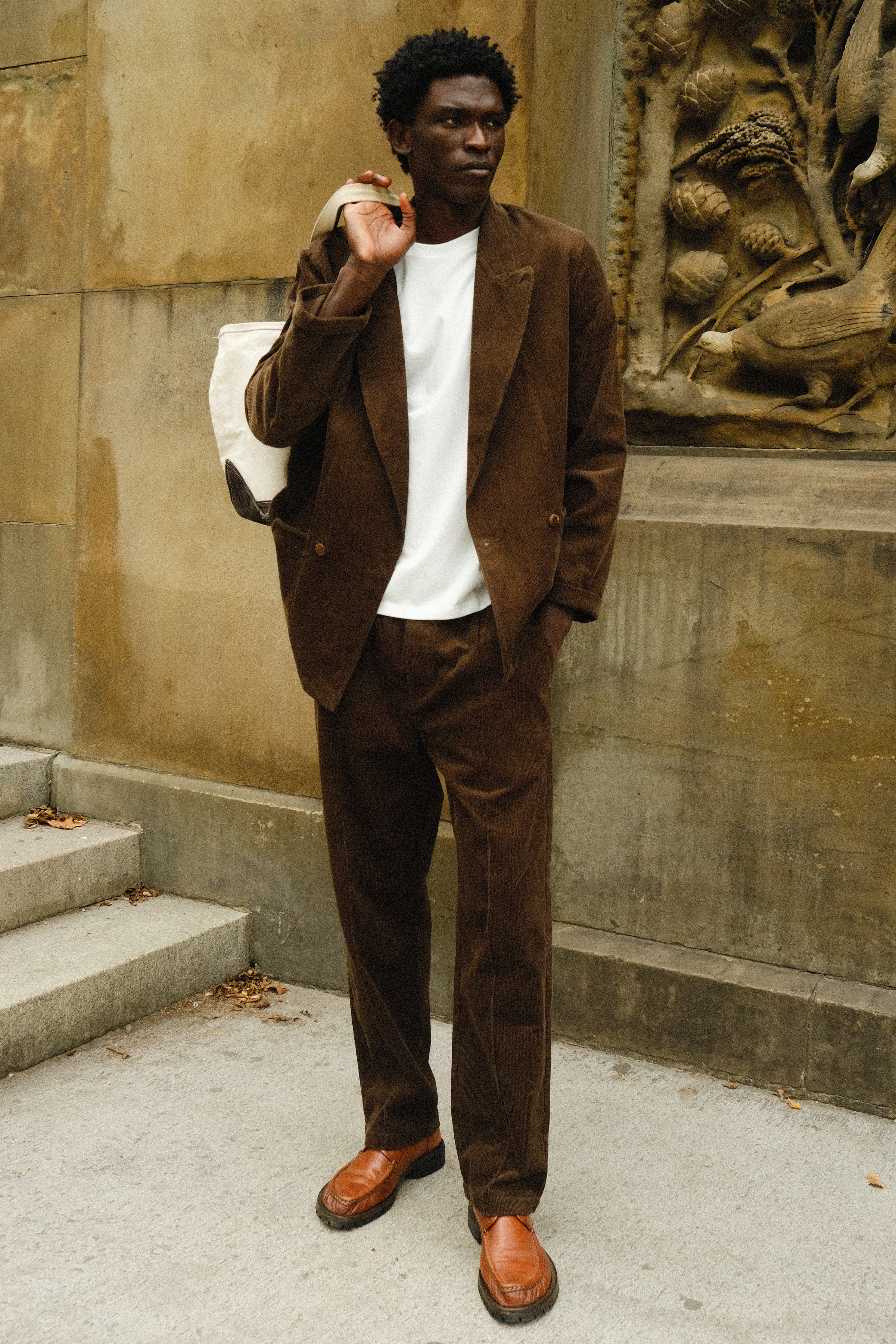 Man wearing a brown corduroy jacket and matching pants with a white T-shirt, brown loafers, and a white canvas tote bag slung over his shoulder.