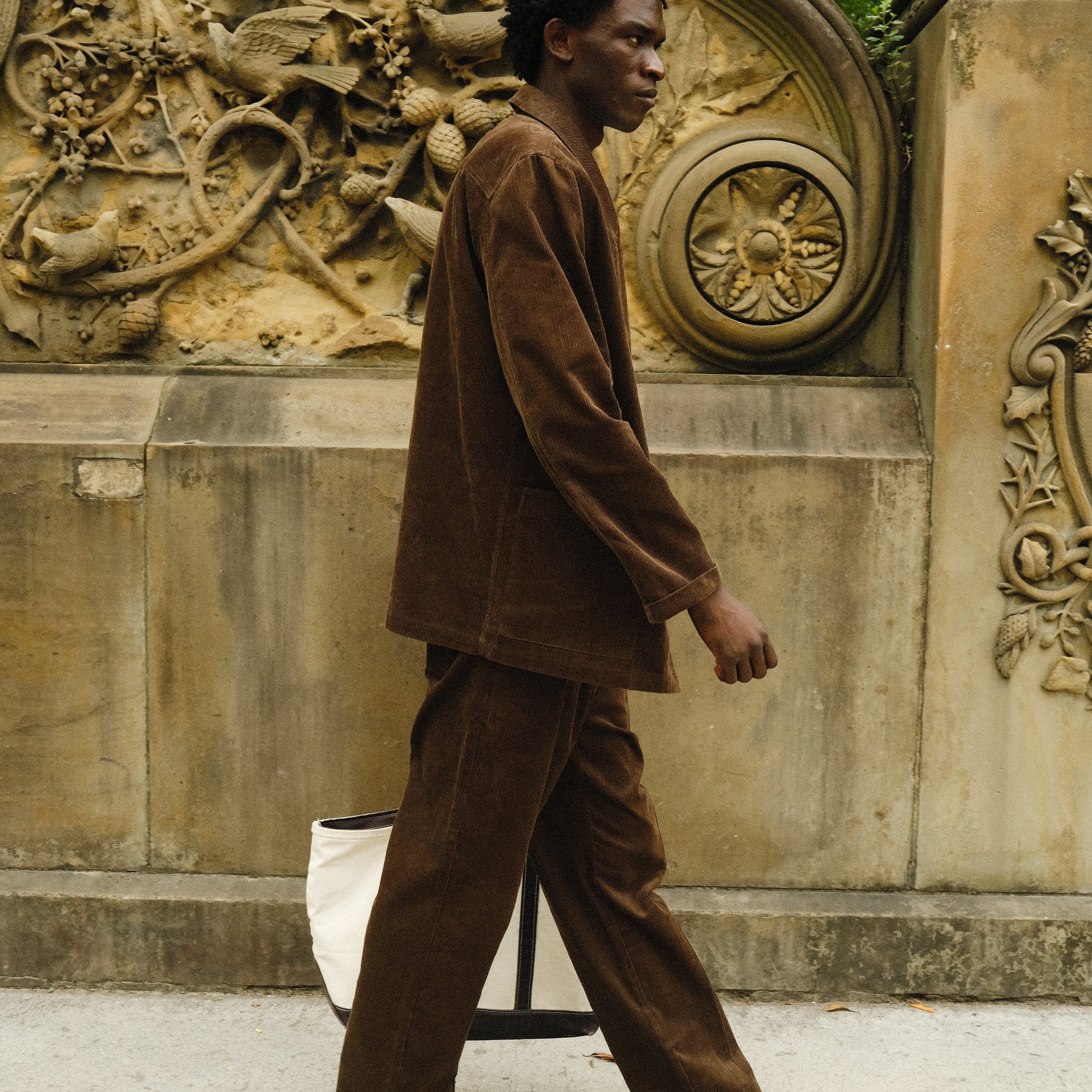 Man walking in a brown corduroy jacket and matching pants, paired with brown loafers and carrying a white canvas tote bag.