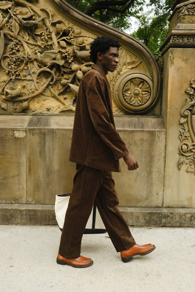 Man walking in a brown corduroy jacket and matching pants, paired with brown loafers and carrying a white canvas tote bag.