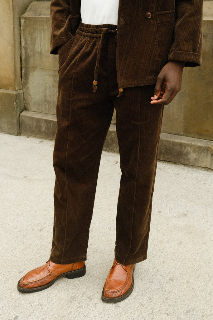 Close-up of a person wearing brown corduroy drawstring pants with a matching jacket, paired with a white shirt and brown loafers.