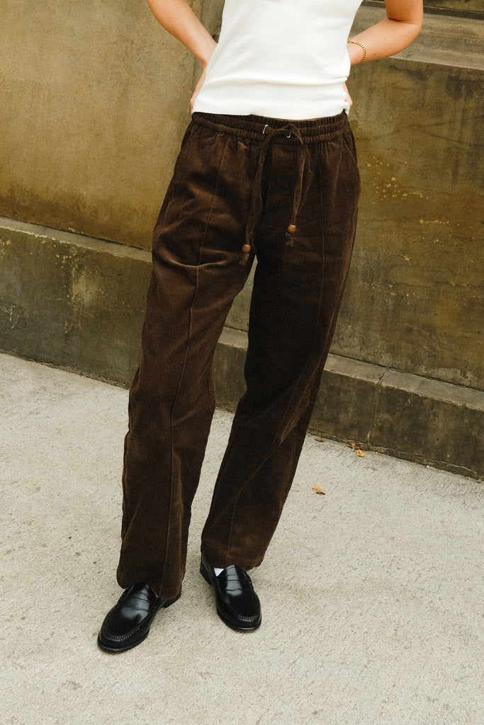 Close-up of a person wearing brown corduroy drawstring pants with a white sleeveless top and black loafers.