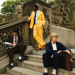 Three people wearing corduroy suits: one in brown with a striped tie, one in bright yellow with a blue shirt and black tie, and one in navy blue with a cream sweater and socks. They are seated and standing on outdoor stone steps.