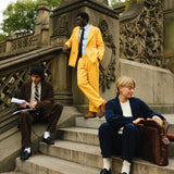 Three people wearing corduroy suits: one in brown with a striped tie, one in bright yellow with a blue shirt and black tie, and one in navy blue with a cream sweater and socks. They are seated and standing on outdoor stone steps.