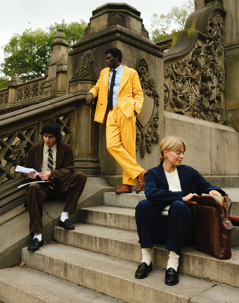 Three people wearing corduroy suits: one in brown with a striped tie, one in bright yellow with a blue shirt and black tie, and one in navy blue with a cream sweater and socks. They are seated and standing on outdoor stone steps.