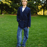 Woman standing on grass wearing a navy blue corduroy jacket over a white shirt, paired with loose-fitting blue jeans and black shoes. 