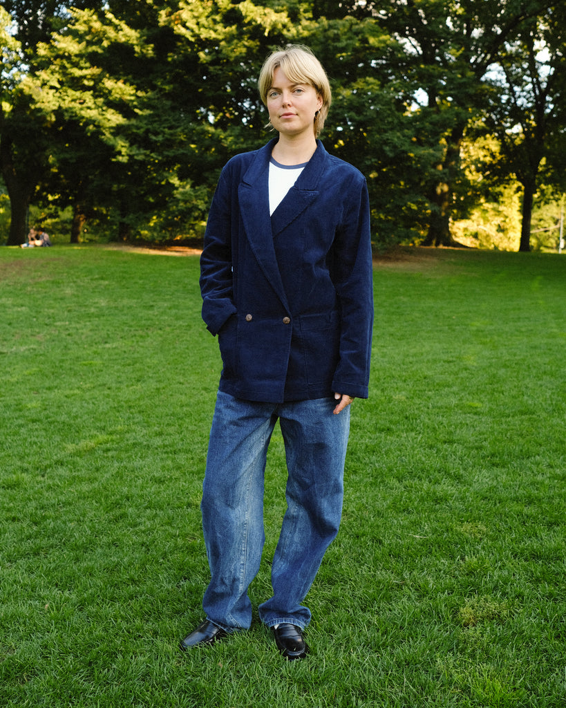 Woman standing on grass wearing a navy blue corduroy jacket over a white shirt, paired with loose-fitting blue jeans and black shoes. 