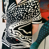 Artist Julian MacMillan wearing black short sleeve cabana shirt with cream chain-stitch embroidery in a loon-inspired dotted and wave pattern, button front.