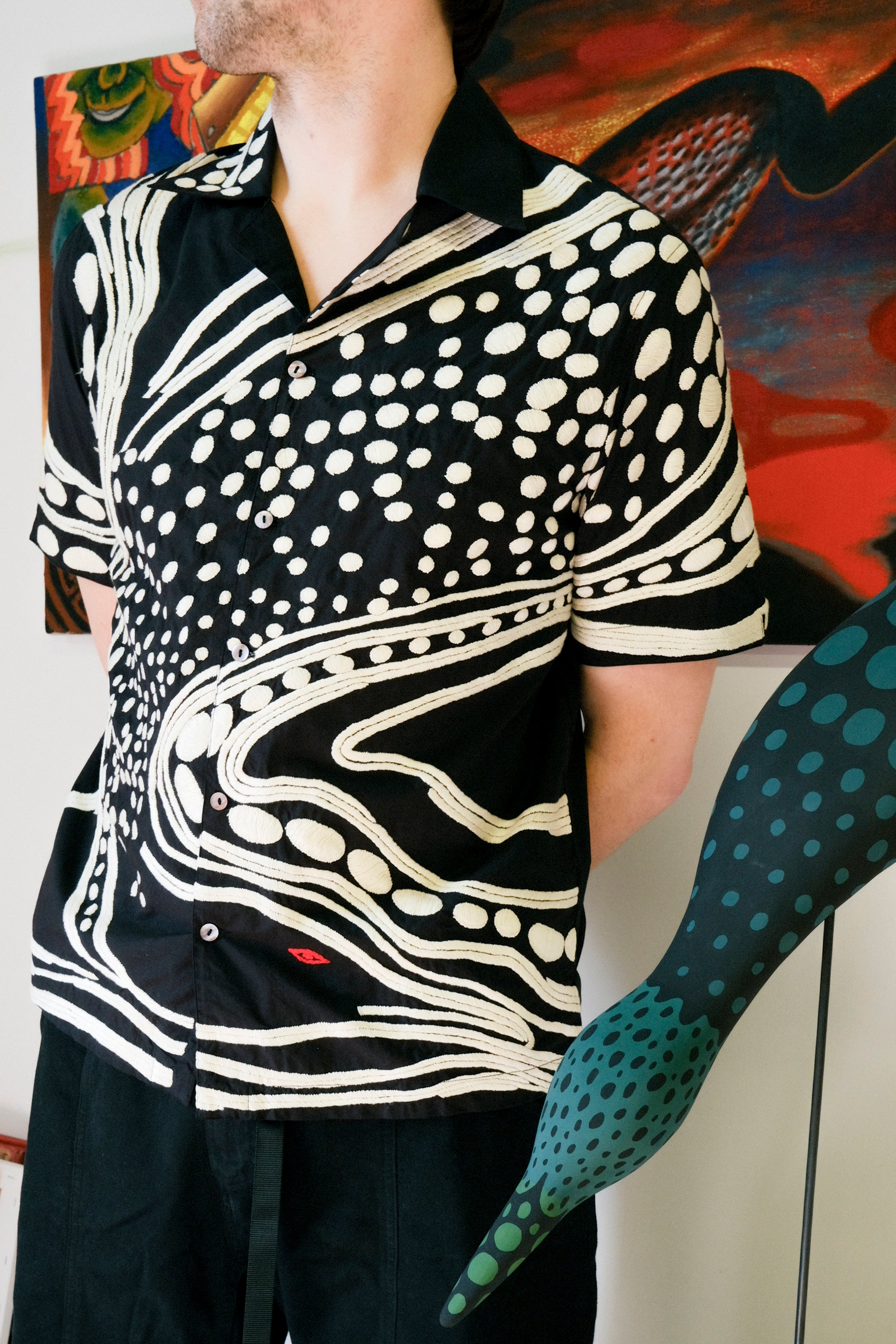 Artist Julian MacMillan wearing black short sleeve cabana shirt with cream chain-stitch embroidery in a loon-inspired dotted and wave pattern, button front.
