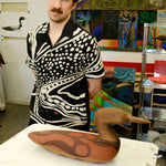 Artist, Julian MacMillan wearing black cabana shirt with cream loon-inspired chain-stitch embroidery, standing beside a carved loon sculpture.