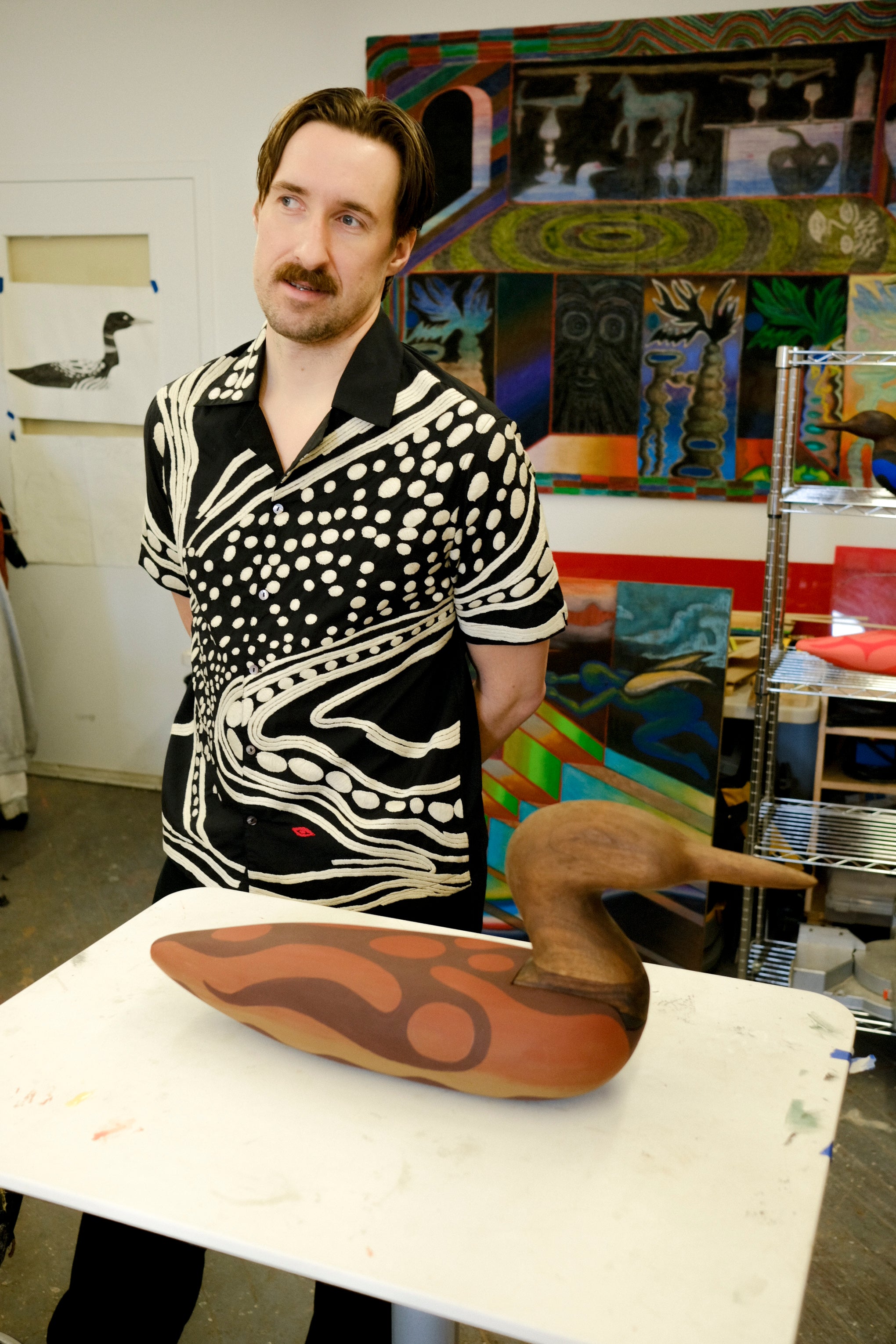 Artist, Julian MacMillan wearing black cabana shirt with cream loon-inspired chain-stitch embroidery, standing beside a carved loon sculpture.