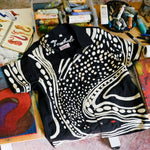 Black short sleeve cabana shirt with cream chain-stitch embroidery in a loon-inspired dotted and wave pattern, laid on a work table with art supplies.