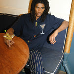 Image of male model seated at a restaurant, wearing the 'Extra Dirty' ~ Relaxed Fit and our Striped Easy Going Pants, and holding a dirty martini with olives in his right hand.