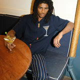 Image of male model seated at a restaurant, wearing the 'Extra Dirty' ~ Relaxed Fit and our Striped Easy Going Pants, and holding a dirty martini with olives in his right hand.