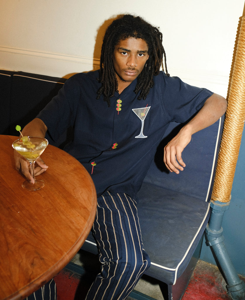 Image of male model seated at a restaurant, wearing the 'Extra Dirty' ~ Relaxed Fit and our Striped Easy Going Pants, and holding a dirty martini with olives in his right hand.