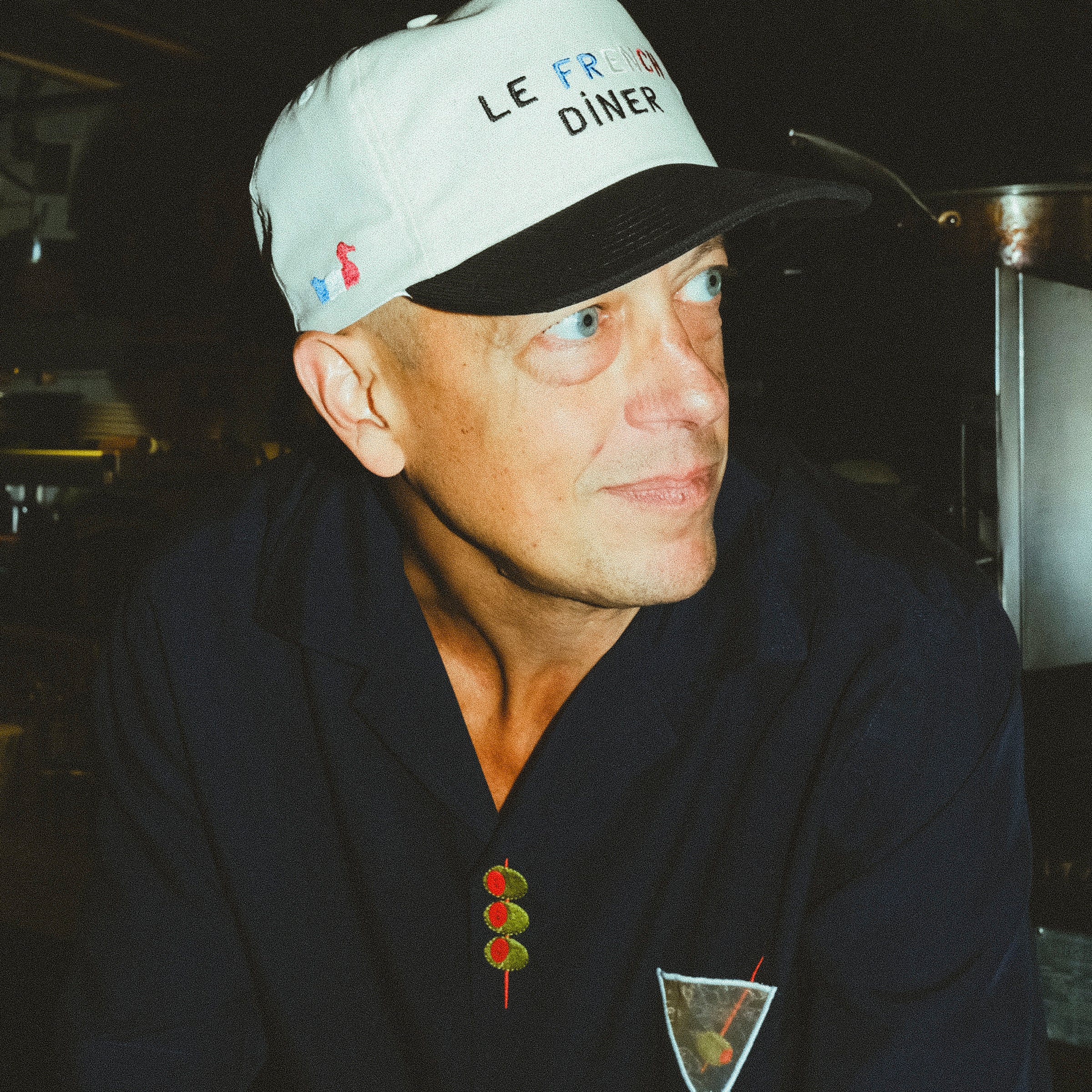 Pierre Moulin, owner of Le French Diner, is pictured wearing the 'Le French Diner' Souvenir Cap. He is also dressed in a navy shirt with embroidered designs of a martini glass and three olives on a skewer.