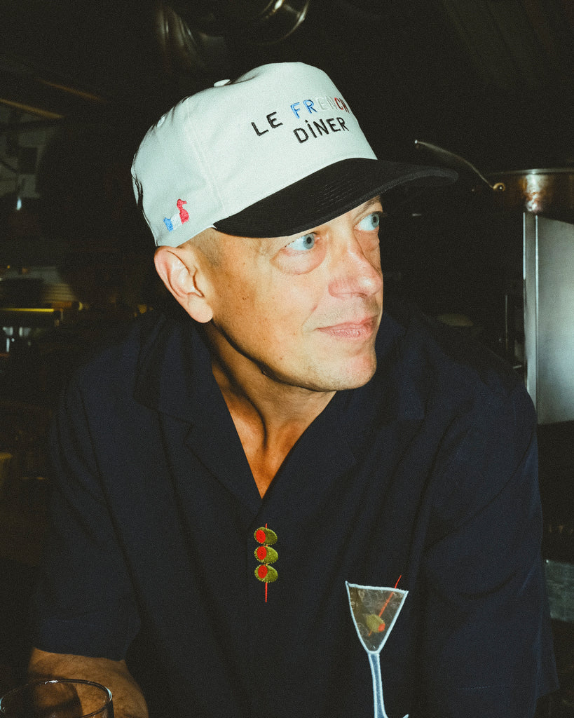 Pierre Moulin, owner of Le French Diner, is pictured wearing the 'Le French Diner' Souvenir Cap. He is also dressed in a navy shirt with embroidered designs of a martini glass and three olives on a skewer.