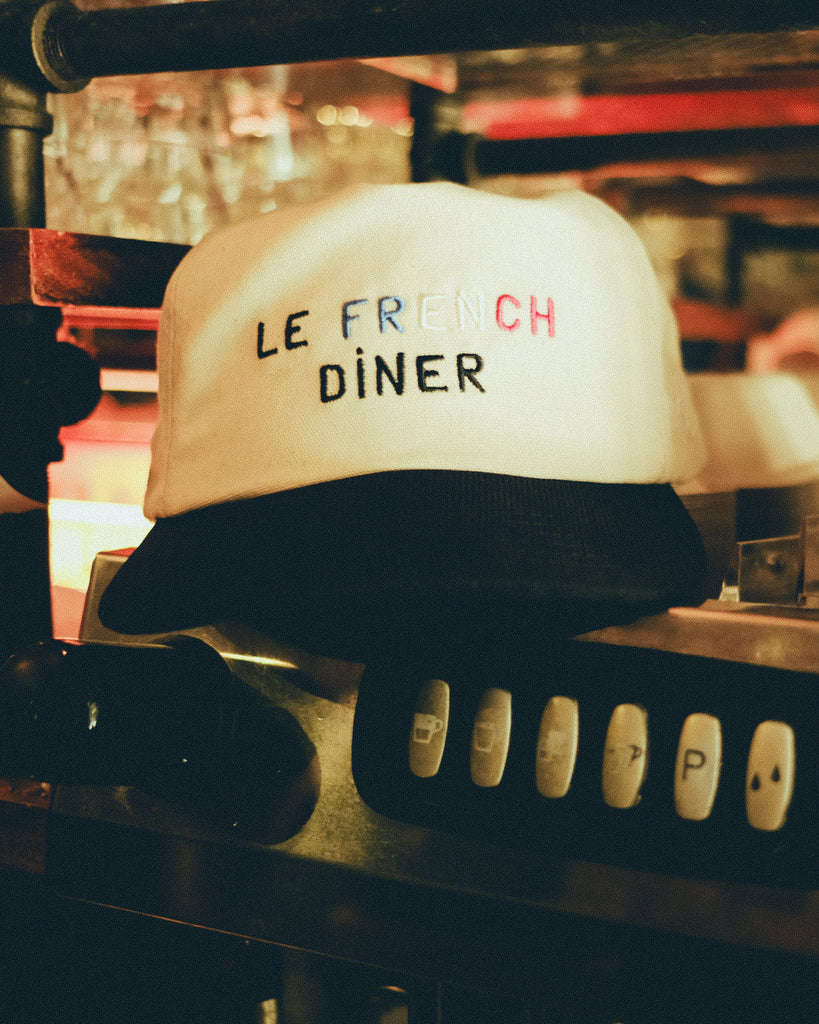 A beige canvas baseball cap with a black brim, embroidered with the words “LE FRENCH DINER” in black and tricolor letters. The cap rests on a metal espresso machine.