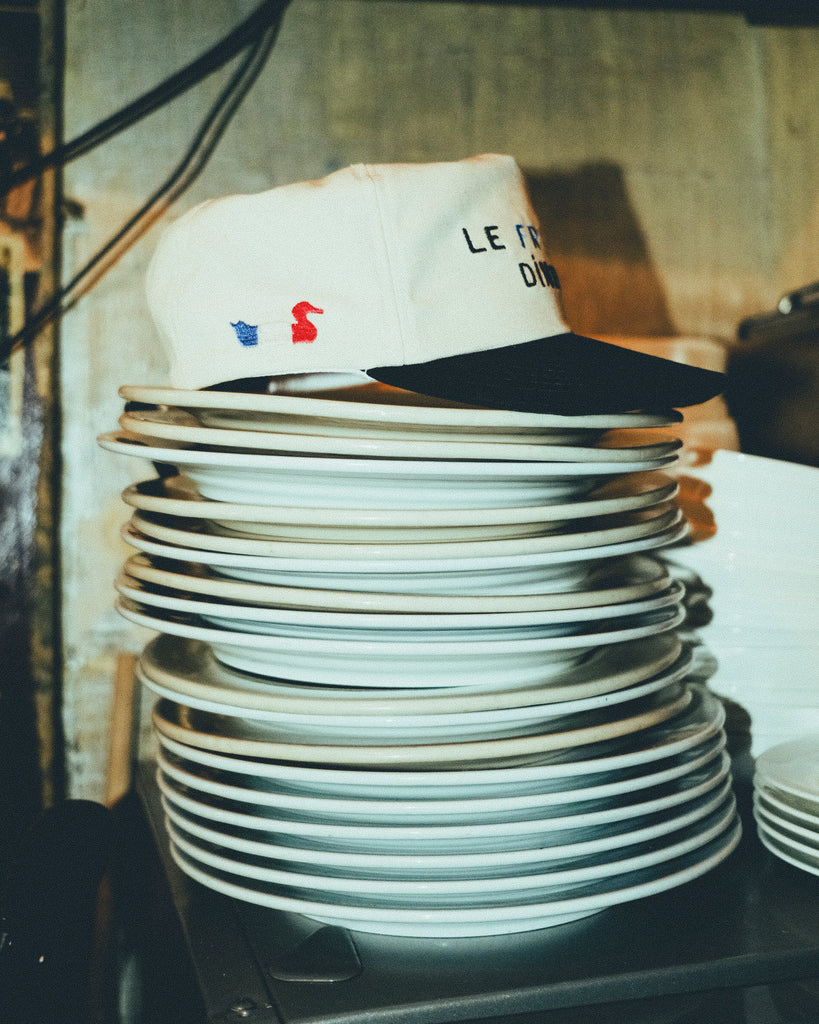 A beige canvas baseball cap with a black brim, embroidered with the words “LE FRENCH DINER.” The cap is resting on top of a tall stack of white plates in a kitchen.