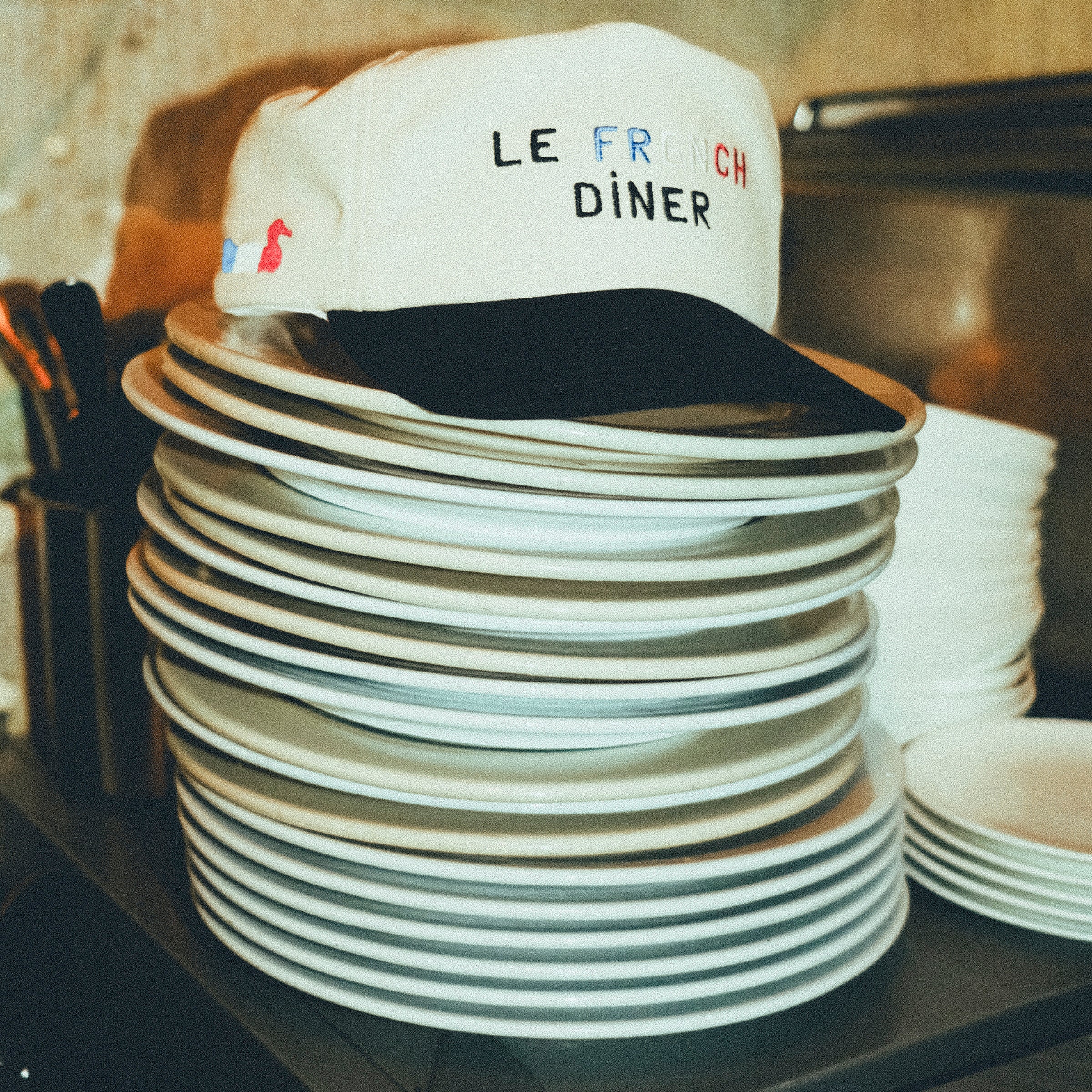 A beige canvas baseball cap with a black brim, embroidered with the words “LE FRENCH DINER.” The cap is resting on top of a tall stack of white plates in a kitchen.