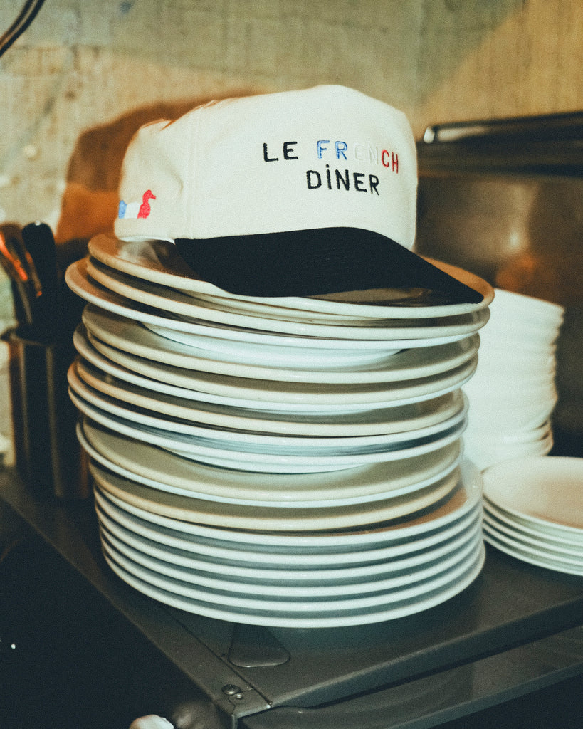A beige canvas baseball cap with a black brim, embroidered with the words “LE FRENCH DINER.” The cap is resting on top of a tall stack of white plates in a kitchen.
